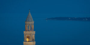 A serene image of Igrane’s iconic 18th-century church tower illuminated at dusk, set against the calm blue Adriatic Sea in the background.