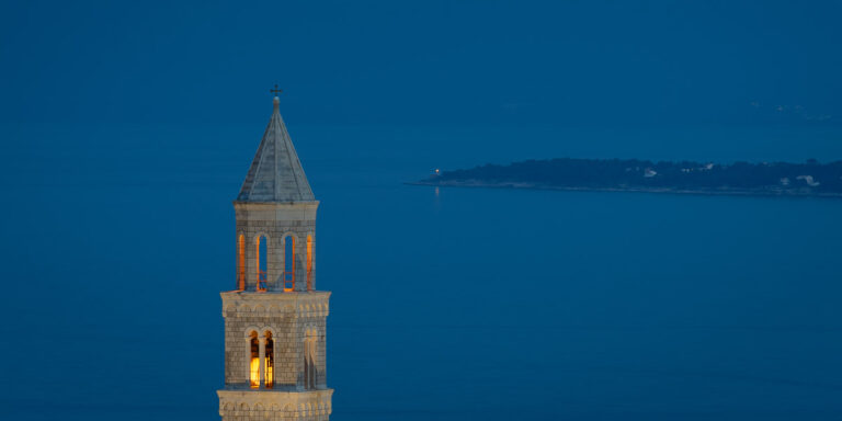 A serene image of Igrane’s iconic 18th-century church tower illuminated at dusk, set against the calm blue Adriatic Sea in the background.